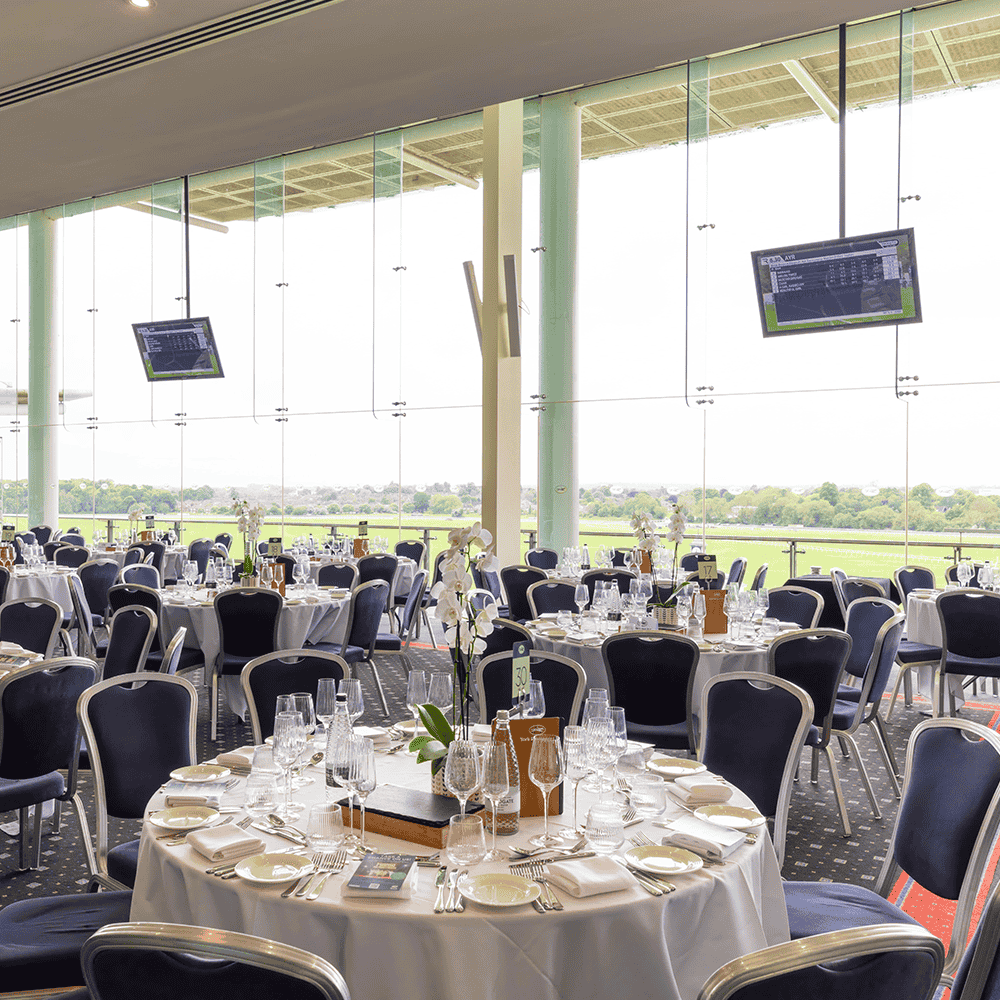 Banquet-style dining setup at Legends Global with round tables, glassware and floral centrepieces, overlooking a large window view of the racecourse.. Legends Global - 1000 x 1000 px