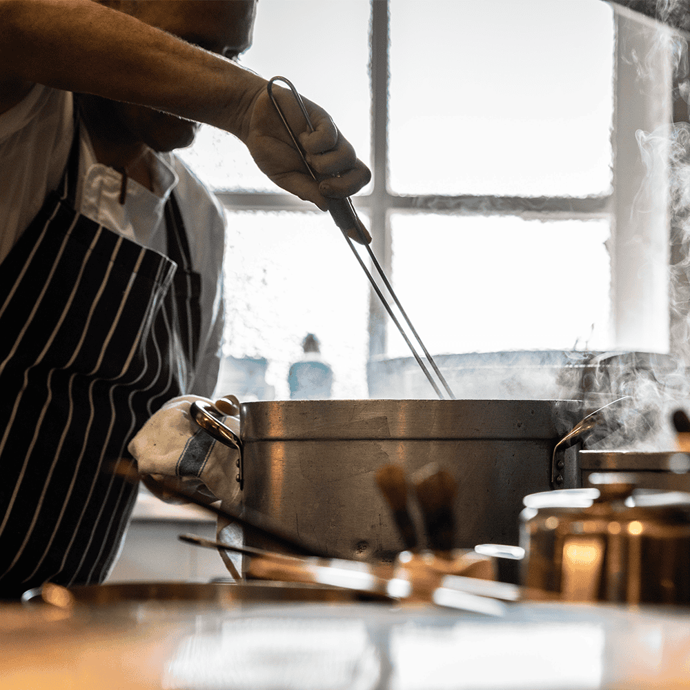 Chef preparing food in kitchen with steaming pot and utensils, behind-the-scenes catering at Legends Global. Legends Global - 1000 x 1000 px