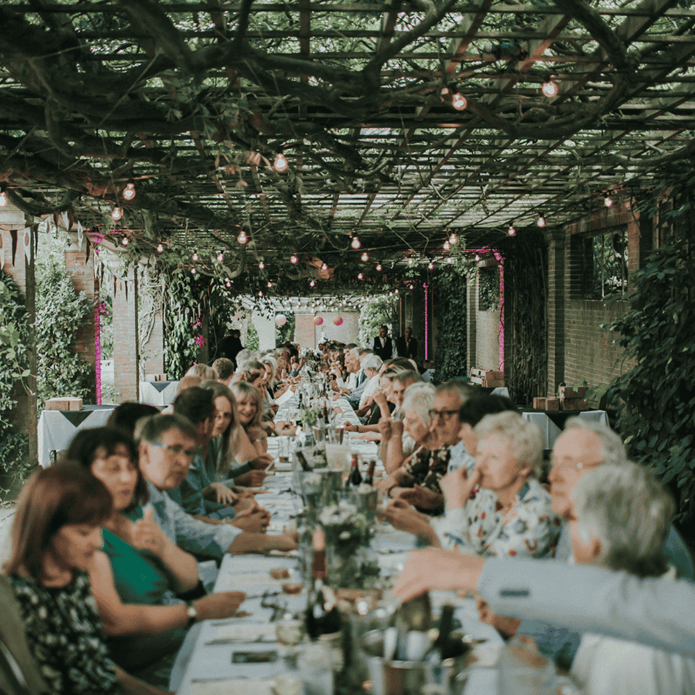 Long communal dining table under a covered terrace with hanging lights and greenery, guests seated and enjoying a meal together. Legends Global - 1000 x 1000 px